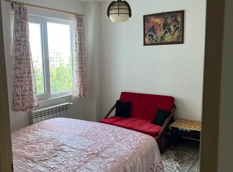 تخت خواب با روتختی صورتی و مبل قرمز رنگ با پنجره رو به محوطه سرسبز اتاق خواب آپارتمان در زرگر