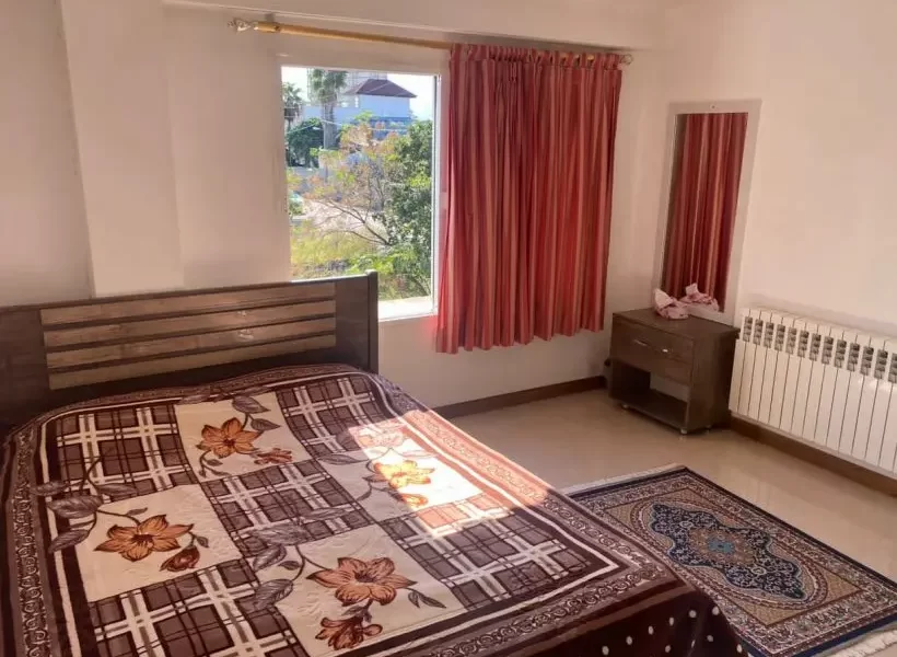 تخت خواب با پتوی قهوهای و پرده ی نارنجی پنجره ی اتاق خواب آپارتمان در پل سفید